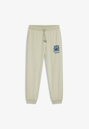 Jasno beżowe dresy z elastycznym pasem i mankietami, z niebieskim graficznym logo na lewej udzie. Gładka faktura materiału.