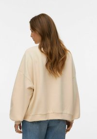 Beežist pehme tekstuuriga suurte õlgadega dressipluus, millel on soonikserv, kombineeritud siniste teksadega. Omaduseks on ümara kaelusega lõige.