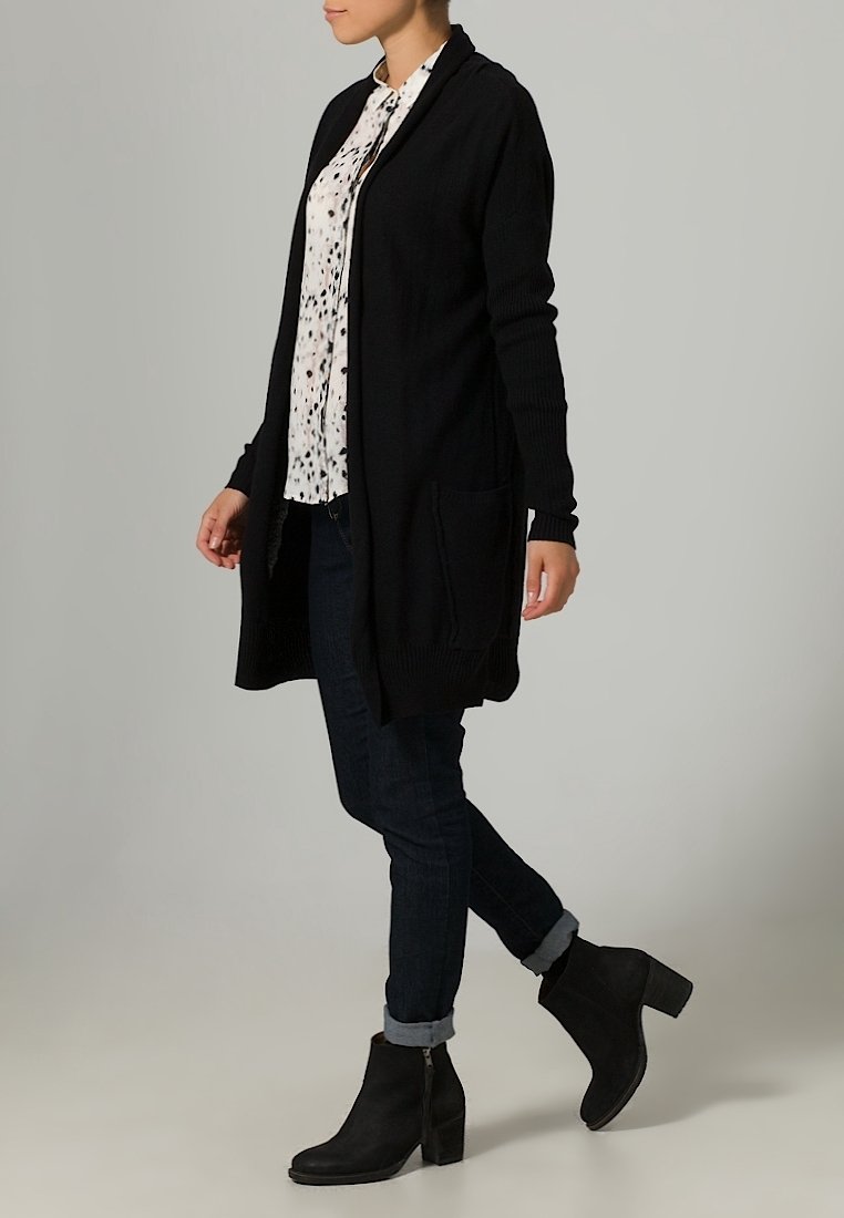 Donna che cammina, vestita con un cardigan lungo nero, una blusa bianca a fantasia, jeans scuri arrotolati e stivaletti neri con tacco, su uno sfondo neutro.