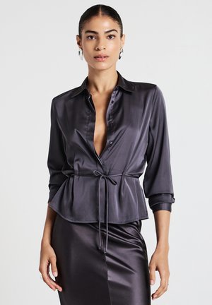 Donna che indossa una blusa di raso scuro con un nodo frontale e una gonna di raso abbinata, abbinata a grandi orecchini d'argento e capelli raccolti.