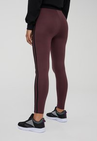 Leggings bordeaux con strisce laterali nere, realizzati in materiale elastico, con una vestibilità affusolata e una superficie liscia. Abbinati a sneaker nere.