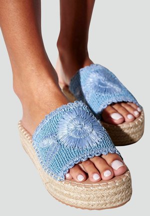 Pieds portant des sandales slides en crochet bleu clair avec des motifs floraux et des semelles compensées épaisses tissées.