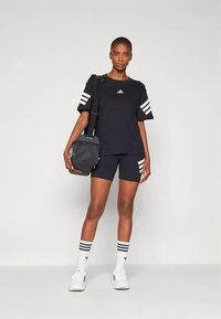 Γυναίκα φοράει μαύρη μπλούζα Adidas και σορτς με λευκές ρίγες, λευκές κάλτσες Adidas, λευκά αθλητικά παπούτσια, και κρατάει μια μαύρη τσάντα ώμου.