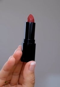 Svart läppstiftflaska med en spetsig, dämpad rosa bullet exponerad. Slät textur, liten rektangulär bas, hålls mellan fingrarna med en ljus nagellack.