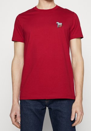 Homme portant un t-shirt rouge à manches courtes avec un petit motif zèbre sur la poitrine gauche et un jean bleu foncé, debout devant un fond uni.