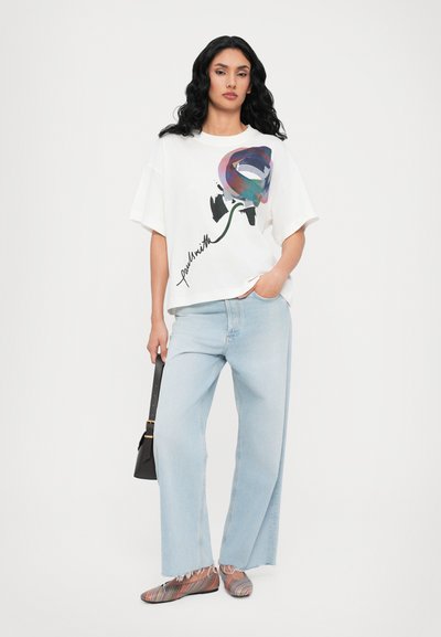 Hvit oversized t-skjorte med fargerik rosegrafikk og svart signatur, lyseblå vide jeans og multifargede stripete sko. Svart veske.
