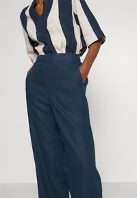 Personne portant un pantalon large bleu marine et une chemise à manches mi-longues avec de larges rayures verticales blanches et bleu foncé, les mains dans les poches.