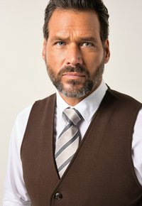 Brauner Weste über einem weißen Hemd, ergänzt durch eine grau gestreifte Krawatte. Die Weste hat einen Knopfverschluss und eine glatte Stoffoberfläche.