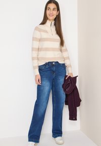 Femme portant un pull rayé beige et blanc, un jean bleu, des baskets blanches, tenant une veste pliée de couleur violet foncé, debout contre des murs blancs.