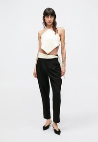 Žena s tetovažama nosi bijelu asimetričnu majicu na jedno rame, crne hlače visokog struka s bijelim remenom i crne štikle sa špicom, stoji.