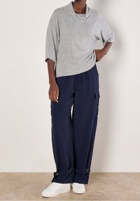 Person trägt ein graues, locker sitzendes Rollkragenoberteil mit marineblauen, weit geschnittenen Cargohosen und weißen Turnschuhen, steht vor einer schlichten Wand.