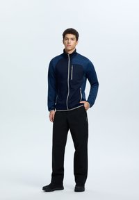 Giacca zip blu navy con pannelli testurizzati di un blu più chiaro, dotata di tasche laterali e pantaloni neri a taglio dritto con una leggera lucentezza.