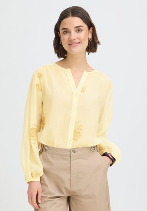 Rövid barna hajú nő halványsárga hímzett blúzban és bézs magasderekú nadrágban, mosolyogva, egyik kezét a zsebében tartva.