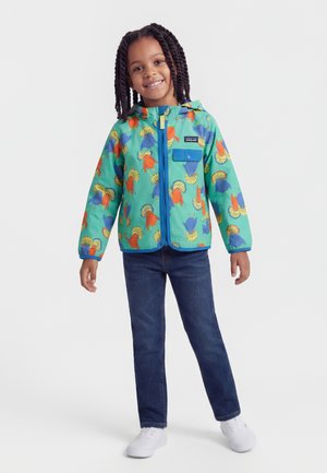 Smilende barn med fletninger, iført en turkis jakke med farverige fugleprint, blå jeans og hvide sneakers, stående med armene let ud til siden.