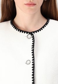 Veste texturée blanche avec des détails en dentelle noire, ornée de boutons en argent avec des accents en perles. Gros plan sur le col et les détails avant.