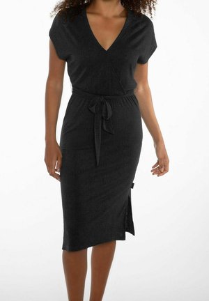 Femme portant une robe portefeuille texturée noire avec manches courtes, ceinture à la taille et fente latérale, debout devant un fond blanc.