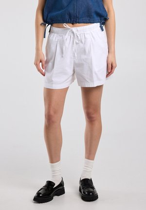 Vrouw draagt witte shorts met trekkoord, witte sokken, zwarte loafers en een kort blauw spijkerjasje, staand tegen een effen achtergrond.