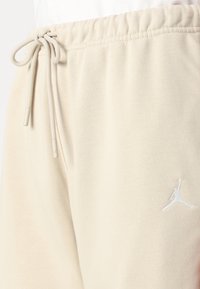 Beige bomulls joggebukser har en elastisk midje med snor og en hvit brodert logo på den nedre delen av benet. Glatt tekstur.