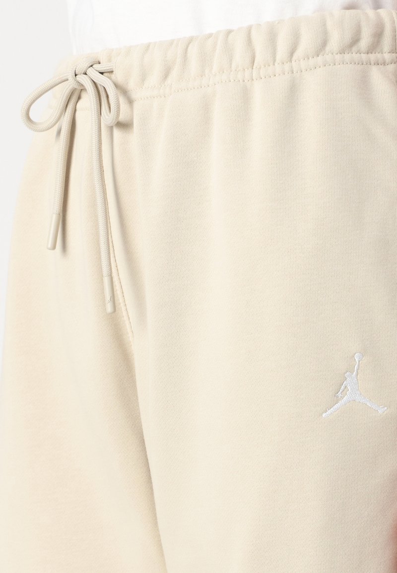 Beige Baumwoll-Sweathosen mit einem elastischen Bund und einem Kordelzug, sowie einem weißen gestickten Logo am unteren Bein. Glatte Textur.