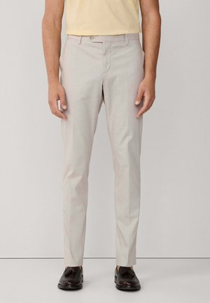 Man wearing light beige slim fit trousers, yellow shirt tucked in, and dark brown leather tassel loafers standing against plain background.