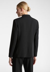 Blazer nero su misura con un design strutturato, texture liscia e uno spacco singolo sul retro, con colletto a pistagna e maniche lunghe.