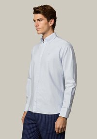 Jeune homme portant une chemise à boutons bleu clair et un pantalon bleu marine à fines rayures, debout devant un fond beige uni.