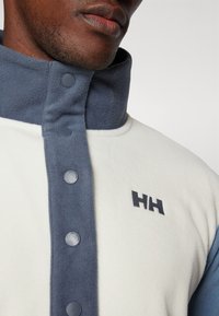 Pullover in fleece con colletto alto, caratterizzato da un corpo bianco, accenti blu scuro, chiusure a bottone e un logo ricamato sul petto.