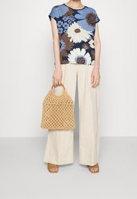 Top a maniche corte a stampa floreale in navy, beige e blu, abbinato a pantaloni larghi beige. Tiene in mano una borsa intrecciata color tan con un manico in legno.