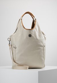 Beige tekstureret tote taske med brune håndtag, metal logo på fronten, sidelommer og en justerbar skulderrem på en hvid pedestal.