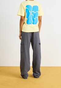T-shirt en coton jaune avec un grand motif de fleur bleue dans le dos, associé à un pantalon cargo gris ample avec des poches latérales.