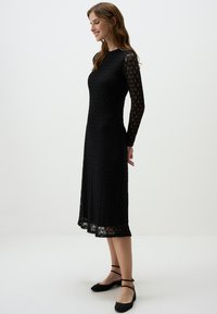 Schwarzes Spitzenkleid mit langen, transparenten Ärmeln, A-Linien-Silhouette, strukturiertem Stoff und knieumspielendem Saum; kombiniert mit flachen schwarzen Riemchenschuhen.