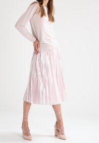 Ljusrosa, långärmad tröja i kombination med en plisserad, glänsande midikjol. Modellen bär beige remsandaler med hög klack. Jämn textur genom hela plagget.