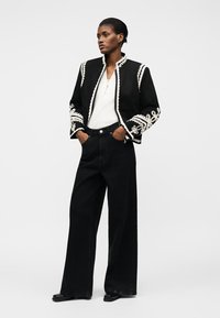 Γυναίκα που στέκεται με τα χέρια στις τσέπες, φοράει μαύρο κεντητό σακάκι, λευκό πουκάμισο, φαρδιά μαύρη παντελόνα και μαύρα παπούτσια.