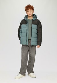 Gepolsterte Jacke mit einem zweifarbigen Design in Türkis und Schwarz, ausgestattet mit einem Reißverschluss, einer Kapuze und gestepptem Material. Getragen über einem grünen Oberteil und grauen Hosen.