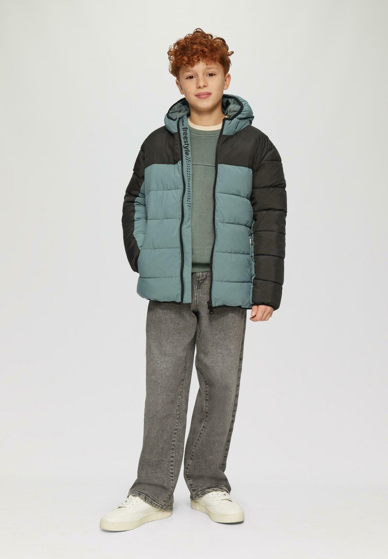 Gepolsterte Jacke mit einem zweifarbigen Design in Türkis und Schwarz, ausgestattet mit einem Reißverschluss, einer Kapuze und gestepptem Material. Getragen über einem grünen Oberteil und grauen Hosen.