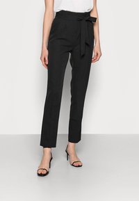 Zwarte op maat gemaakte broeken met een hoge taille, strikceintuur, zijzakken en scherpe plooien. Gecombineerd met zwarte hoge hakken met banden.