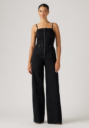 Vrouw staand, gekleed in een zwarte mouwloze jumpsuit met een rits aan de voorkant en broek met wijde pijpen tegen een effen lichte achtergrond.