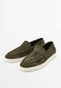 Zamszowe buty slip-on w ciemnozielonym kolorze o gładkiej teksturze, z zaokrąglonym noskiem i białymi gumowymi podeszwami. Dekoracyjne przeszycia wzdłuż szwów.