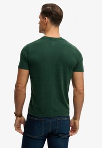 Grøn bomuldst-shirt med rund halsudskæring, korte ærmer og et mærke på øverste ryg. Brugt med mørkeblå jeans. Glat tekstur.