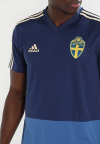 Marinblå sporttröja i syntetmaterial med guldaccenter, försedd med Adidas-logotypen och en svensk emblem på bröstet.