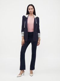 Femme debout portant un pantalon bleu marine et un blazer avec des manches de sweat à capuche gris, une chemise rose et des talons blancs à bout pointu sur fond blanc.