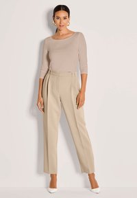 Vrouw in een beige, nauwsluitend langmouwtop, hoge taille plissé beige broeken, witte hakken en gouden ringoorbellen, staand tegen een effen muur.