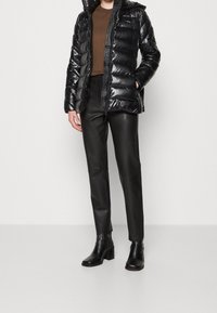 Persona vestida con una blusa marrón, una chaqueta de plumas negra y brillante, pantalones de cuero negros y botines negros con tacón, de pie contra un fondo sencillo.