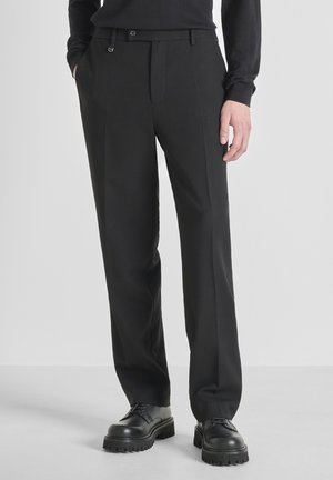 Pantalones negros con un diseño a medida, con frente plano, pasadores para cinturón y bolsillos laterales, hechos de una tela suave, combinados con unos zapatos negros gruesos.