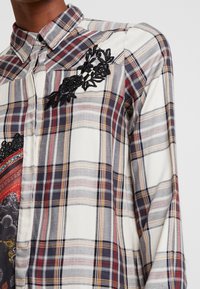 Camisa de cuadros en blanco, azul marino y burdeos con cierre de botones en la parte delantera. Presenta bordados florales negros en el pecho y una tela suave y texturizada.