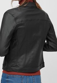 Chaqueta de cuero negra con un diseño ajustado, costuras visibles y cuello alto. Llevada sobre un suéter color óxido y jeans de mezclilla.