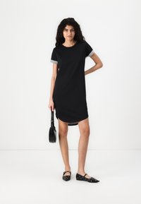 Robe noire à manches courtes avec un revers gris, col rond et ourlet arrondi. Chaussures plates noires avec des accents circulaires et un petit sac à main.