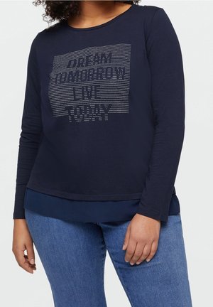 Kobieta w granatowej bluzce z długim rękawem z napisem z kryształków "DREAM TOMORROW LIVE TODAY" i niebieskich dżinsach na białym tle.
