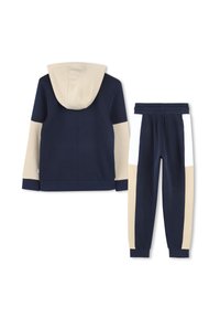 Marineblauwe en beige hoodie met een voorzak en een capuchon, gedragen met marineblauwe sweatpants met beige en witte zijpanelen. Zachte gebreide stof.