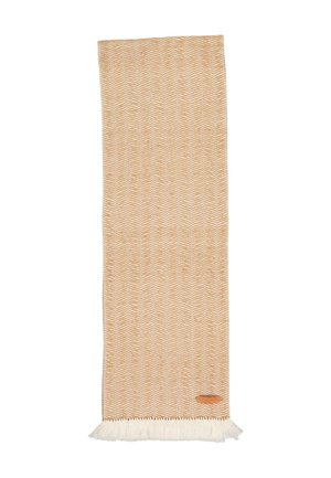 Tapis rectangulaire beige tissé avec un motif zigzag blanc, bords frangés et petite étiquette en cuir marron dans le coin inférieur droit.